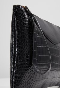 Sac à main en cuir noir avec texture crocodile, rabat incurvé et sangle unique, présenté en vue latérale inclinée sur un fond neutre.
