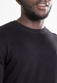 Czarny dziergany sweter z okrągłym dekoltem, o gładkiej fakturze i ściągaczach przy mankietach. Subtelne logo na wysokości klatki piersiowej.