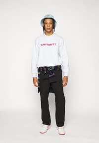 Grauer Sweatshirt mit rotem "carhartt." Logo, schwarze Hose und weiße Turnschuhe mit roten Akzenten. Mehrfarbiger gestreifter Stoff um die Taille gebunden.