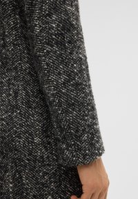 De hand van een persoon ligt naast de gehoekte zwarte en witte herringbone wollen mouw van een jas tegen een effen achtergrond.