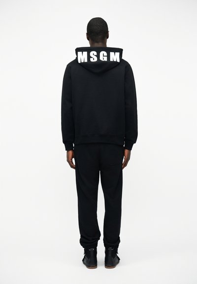 Personne debout, vue de dos, portant un sweat à capuche noir avec un grand texte blanc "MSGM" sur la capuche, un pantalon noir et des chaussures noires.