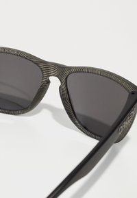Ochelari de soare cu dungi negre și gri, cu lentile închise, având un cadru gros și brațe rezistente, logo pe lateral și un design care se înfășoară.