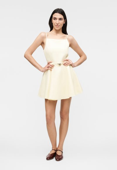 Γυναίκα που φοράει ένα μίνι αμάνικο φόρεμα εκρού με λεπτές τιράντες και ζώνη, σε συνδυασμό με σκούρα καφέ παπούτσια Mary Jane, στέκεται με τα χέρια στη μέση.