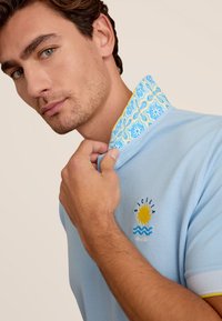 Lichtblauwe polo met een gepatterneerde kraag in geel en blauw, geborduurde "Sicilia" met een zon- en golfontwerp, korte mouwen.