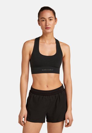 Mujer con un sujetador deportivo negro y shorts deportivos negros, de pie mirando al frente con una expresión neutral sobre un fondo blanco.