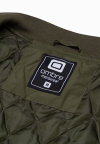 Olivgrön vadderad jacka inuti med en svart etikett som har "ombre menswear" i vit text och storlek "M." Inkluderar en upphängningsögla.