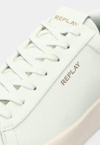 Zapatilla de cuero blanco con cordones planos, un diseño minimalista y un sutil logo en relieve "REPLAY" en dorado en el costado. Textura suave.