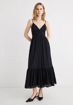 Frau trägt ein ärmelloses schwarzes Maxikleid mit Spaghettiträgern, V-Ausschnitt, geraffter Taille und Volant-Saum, kombiniert mit schwarzen spitzen Schuhen.