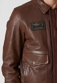 Giacca di pelle marrone con tasca a risvolto e patch ricamato con la scritta "1 Leader" con ali e corona sul petto.