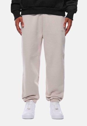 Lys beige joggebukser med avslappet passform, laget av myk stoff. Har en liten brodert logo og ribbestrikkede anklestropp. Brukt med hvite sneakers.