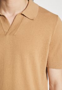 Gros plan d'une personne portant un polo en maille à manches courtes de couleur chameau avec un col en V.