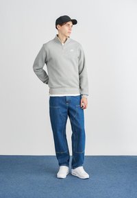 Šedý fleecový svetr na zip, modré džíny a bílé tenisky, doplněné černou kšiltovkou. Jednoduchý design s minimálním označením na tričku.