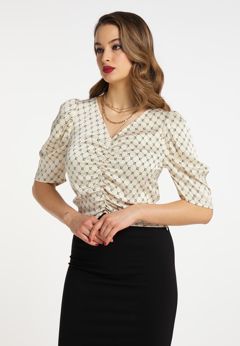 Cremefarvet bluse med korte pufærmer, prydet med sort geometrisk mønster og rynk ved fronten, kombineret med en sort blyantskirt.