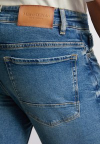 Jeans in denim blu con un'etichetta in pelle marrone cucita sulla vita posteriore, design standard a cinque tasche e dettagli di cucitura a contrasto.