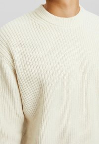 Sweter w kolorze kremowym, wykonany na drutach, o teksturze prążkowanej, z okrągłym dekoltem i luźnym krojem. Widoczne detale szycia na tkaninie.