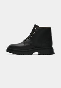 Stivaletto in pelle nera con punta tonda, occhielli dorati e suola texturizzata. Presenta un colletto imbottito e un logo distintivo sul lato.