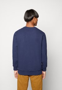 Navy blue crewneck sweatshirt made of soft fabric, featuring long sleeves and ribbed cuffs, worn over mustard-colored pants.