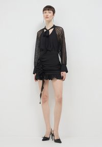 Eine Frau trägt eine schwarze Spitzenbluse mit langen Ärmeln und einer Schleifen-Nelke, einen gerafften Minirock mit Rüschendetails und schwarze High Heels.