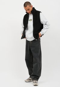 Mustassa sherpa-liivissä on harmaa huppari, jossa on graafinen painatus, yhdistettynä löysän istuvuuden tummansinisiin farkkuihin ja vaaleanruskeisiin lenkkareihin. Yhteenvetona rento tyyli.