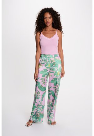 Donna con capelli ricci che indossa una canottiera rosa chiaro e pantaloni a gamba larga con motivi floreali verdi, in piedi davanti a uno sfondo bianco semplice.