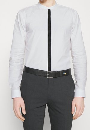 Homme portant une chemise blanche à manches longues avec une bande verticale noire et un pantalon gris foncé maintenu par une ceinture noire avec une boucle rectangulaire.