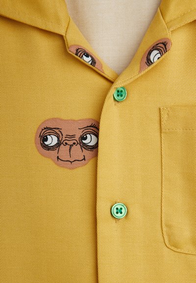 Κίτρινο πουκάμισο φτιαγμένο από υφασμάτινη υφή, με κεντημένο cartoon πρόσωπο και πρ�άσινους κουμπιά στους γιακά και την πατίνα.
