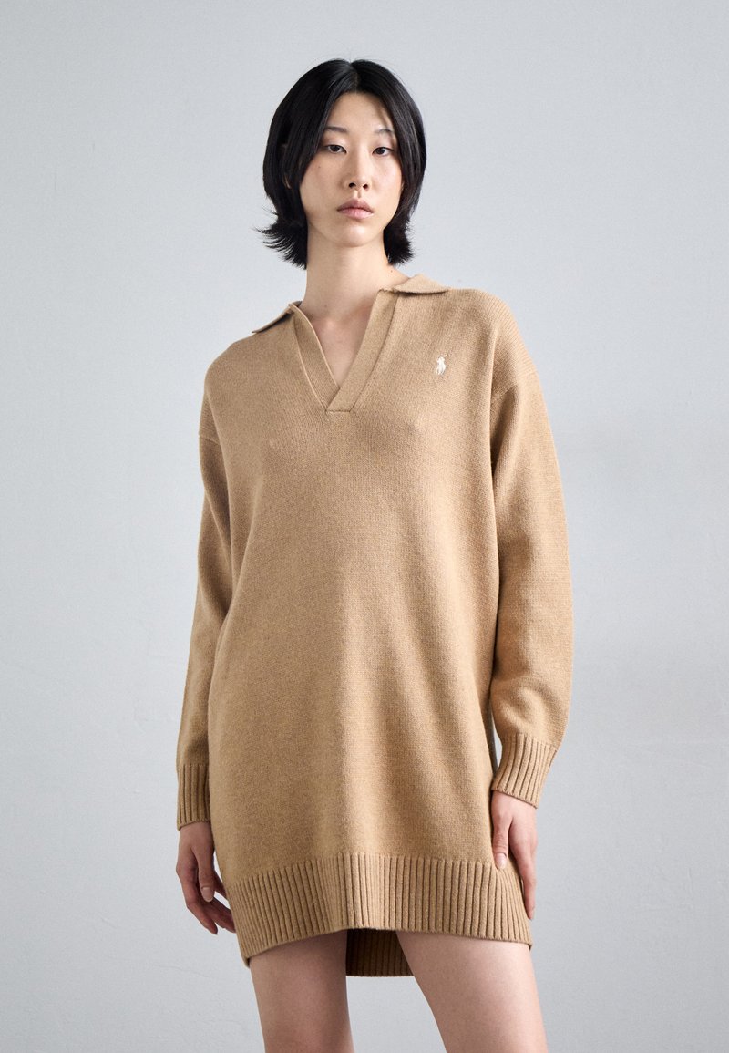 Gebreid beige jurk met V-hals en ribgebreide manchetten. Bevat een klein logo op de borst en heeft een rechte silhoetta. Zachte textuur.