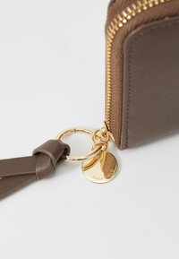 Brun læderpung med guldlås, en nøgleringsstrop og en metalvedhæng med indgraveret "ANNA FIELD." Detaljeret tekstureret overflade synlig.