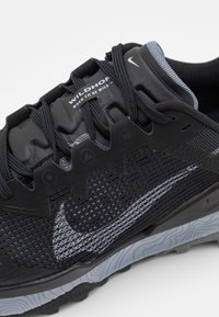 Scarpa sportiva nera realizzata in materiale mesh, con accenti riflettenti e suola grigia testurizzata. Presenta un sistema di allacciatura e dettagli in gomma sui lati.
