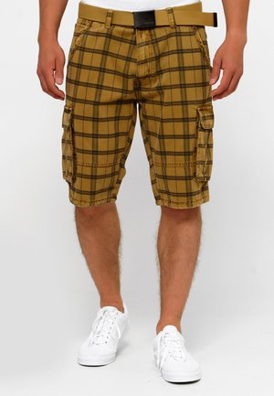 Mann trägt senfgelbe karierte Cargo-Shorts mit Seitentaschen und passendem Gürtel, kombiniert mit weißen Sneakers und einem weißen Hemd.
