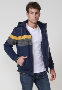 Chaqueta con cremallera de color azul marino con una franja amarilla en contraste, panel de pecho de punto texturizado y capucha forrada. Presenta bolsillos laterales y puños de canalé.