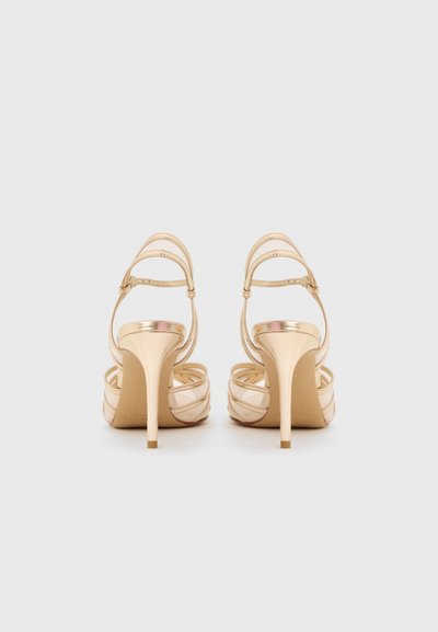 Ζευγάρι γυμνών σανδαλιών με λουριά και ψηλοτάκουνες πέδιλες στιλέτο, φαίνονται από πίσω σε απλό ανοιχτό φόντο.