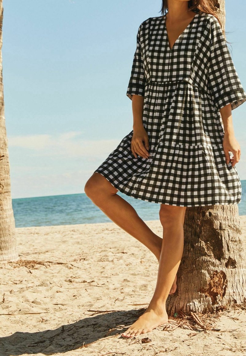 Femme portant une robe à carreaux noire et blanche, debout pieds nus sur le sable, appuyée contre un palmier avec la mer en arrière-plan.