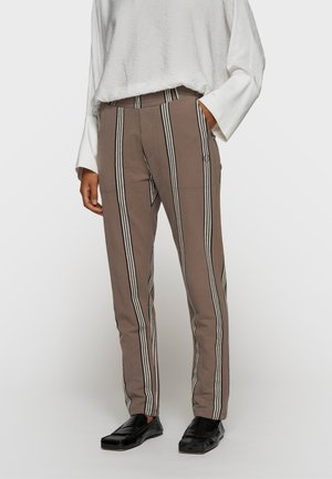Persona che indossa pantaloni marroni con strisce verticali nere e bianche, camicetta bianca a maniche lunghe e mocassini neri, in piedi con una mano in tasca.