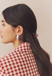 Woman with long dark hair held by a red patterned hair clip, wearing silver teardrop earrings and a red and white checkered shirt.