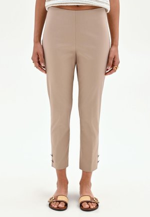 Donna che indossa pantaloni beige corti con dettagli con bottoni vicino alle caviglie e sandali intrecciati, in piedi su sfondo bianco.