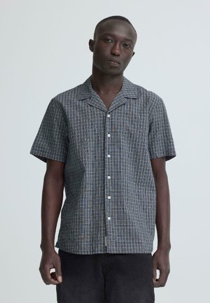 Mann trägt ein graues kariertes Kurzarmhemd mit Knöpfen und dunkle Hosen, steht vor einem hellgrauen Hintergrund.