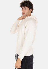 Pullover con cappuccio di colore crema, con polsini e orlo a coste, dotato di una tasca centrale e tessuto strutturato. Modella in posa di lato.