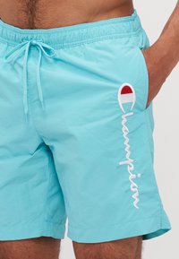 Shorts de bain turquoise avec une taille élastique et un cordon de serrage, présentant du texte et un logo "Champion" brodés en blanc sur le côté.