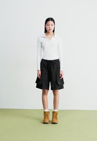 Weißes Langarm-Polohemd, schwarze knielange Shorts mit Taschen und gelb-braune Schnürstiefel. Schlichter Hintergrund und grüner Boden.