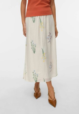 Witte midi-rok met kleurrijke bloemrijke borduursels, gedragen met bruine pumps met puntige neus en een oranje mouwloze top, tegen een effen achtergrond.