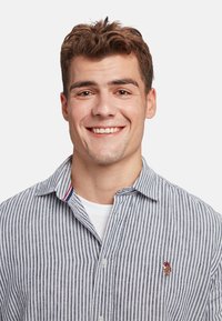 Camicia a righe con bottoni in blu e bianco, caratterizzata da un piccolo accento ricamato sul lato sinistro del petto e un colletto a contrasto con dettagli rossi.