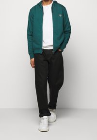 Türkise Zip-Hoodie mit Kapuze, weißes T-Shirt darunter, schwarze Hose und weiße Sneaker. Verfügt über Logodetails auf der Brust.