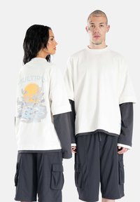 Bavlnené oversized tričko v odtieňoch off-white, s pastelovou grafikou vĺn a slnka na chrbte. V kombinácii s tmavými cargo šortkami.