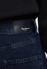 Tamno plave traperice imaju crnu kožnu oznaku s bijelim natpisom "Pepe Jeans LONDON", detaljno šivanje i više džepova.