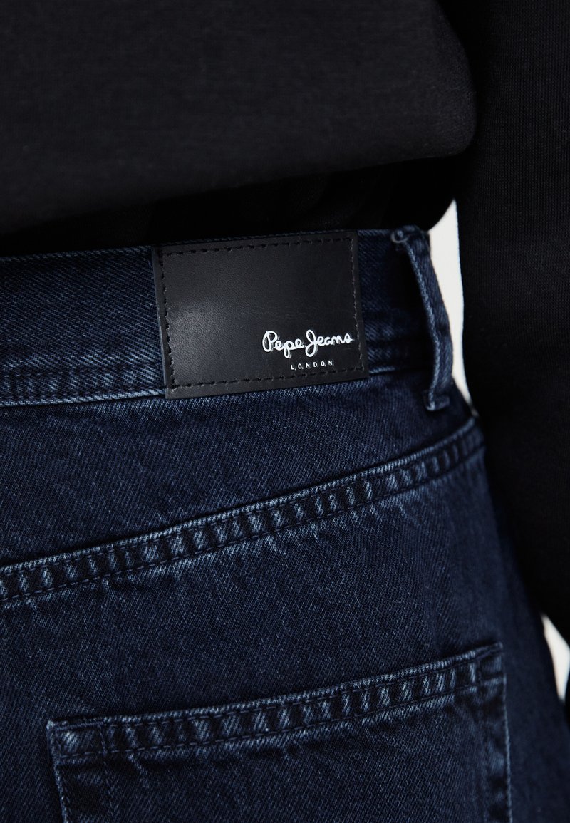 I jeans in denim blu scuro presentano un'etichetta in pelle nera con testo bianco che riporta "Pepe Jeans LONDON", cuciture dettagliate e numerose tasche.