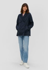 Marineblaues, doppelt geknöpftes Jackett mit zwei vorderen Taschen und großen Knöpfen, kombiniert mit hellblauen Jeans und weißen Sneakers. Glatte Stofftextur.