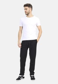 Biała koszulka z okrągłym dekoltem, czarne spodnie dresowe z sznurkiem, czarne sneakersy z białymi paskami, stojący z jedną ręką w kieszeni, jednolite tło.