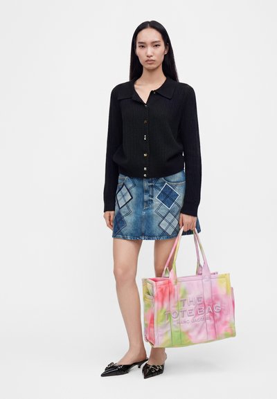 Femme portant un pull noir boutonné et une mini-jupe en jean à motifs, tenant un grand sac fourre-tout tie-dye et portant des chaussures noires pointues.