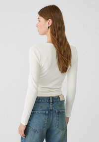 Weißes, geripptes Langarmtop kombiniert mit hoch geschnittenen blauen Denim-Jeans. Die Jeans verfügen über zwei Gesäßtaschen und ein hellbraunes Etikett am Bund.